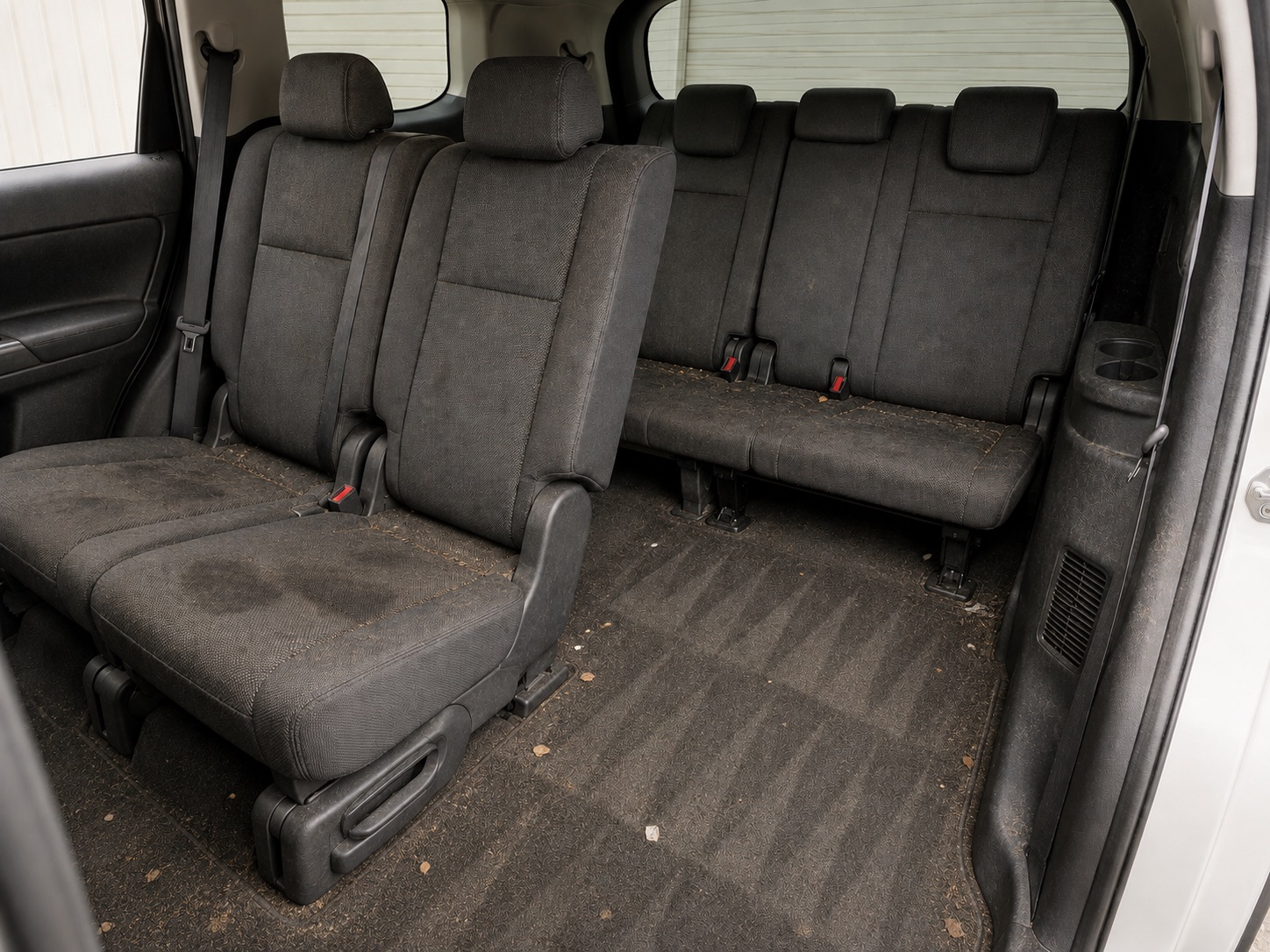 The rear rows of an SUV before cleaning, with crumbs and debris across the seats and matted, soiled carpet