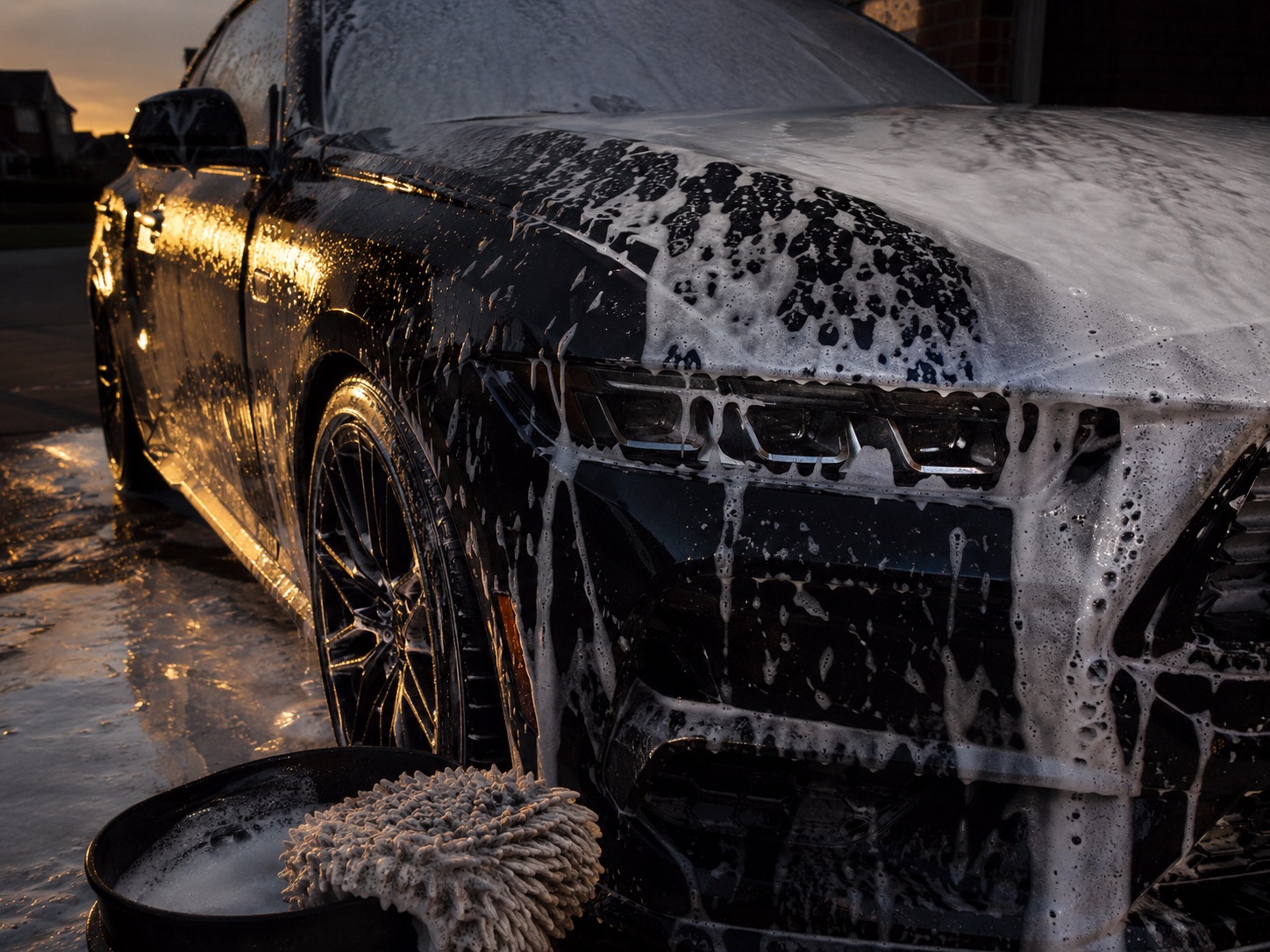 A dark car covered in wash foam on a driveway at sunset, with a wash mitt resting on a bucket in the foreground