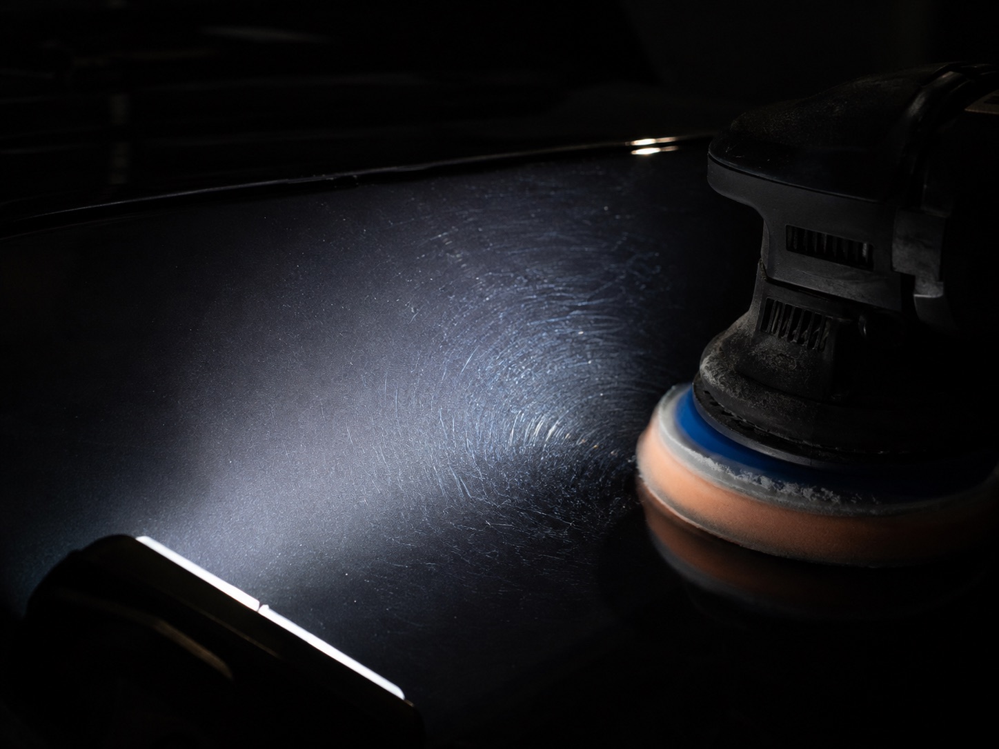 A dual-action polisher resting on a dark panel, with an inspection light raking across the paint to reveal fine swirl marks